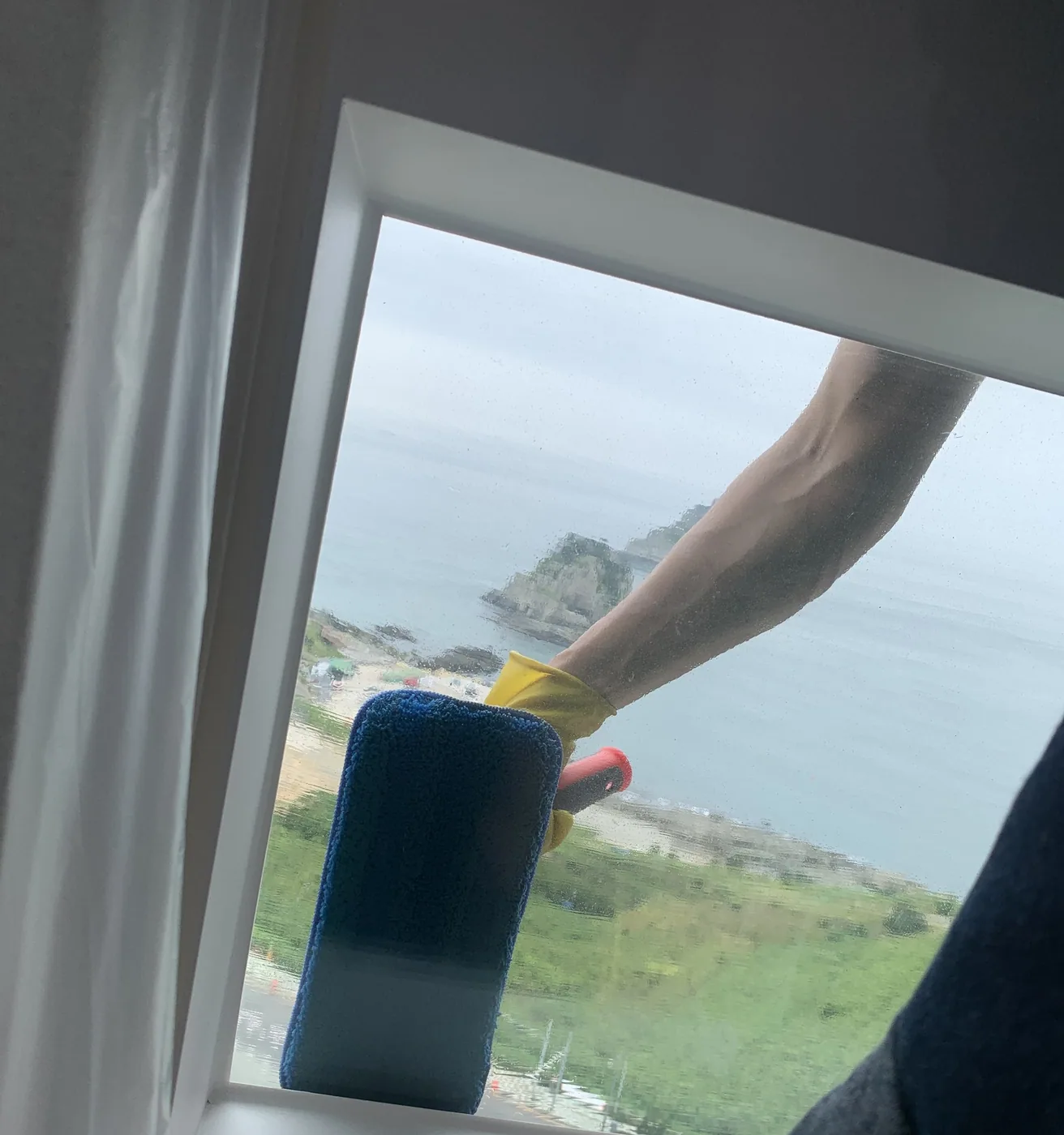고무장갑을 낀 작업자의 손이 워셔를 쥐고 창 유리 가장자리를 문지르는 모습, 창밖으로 바다가 보인다