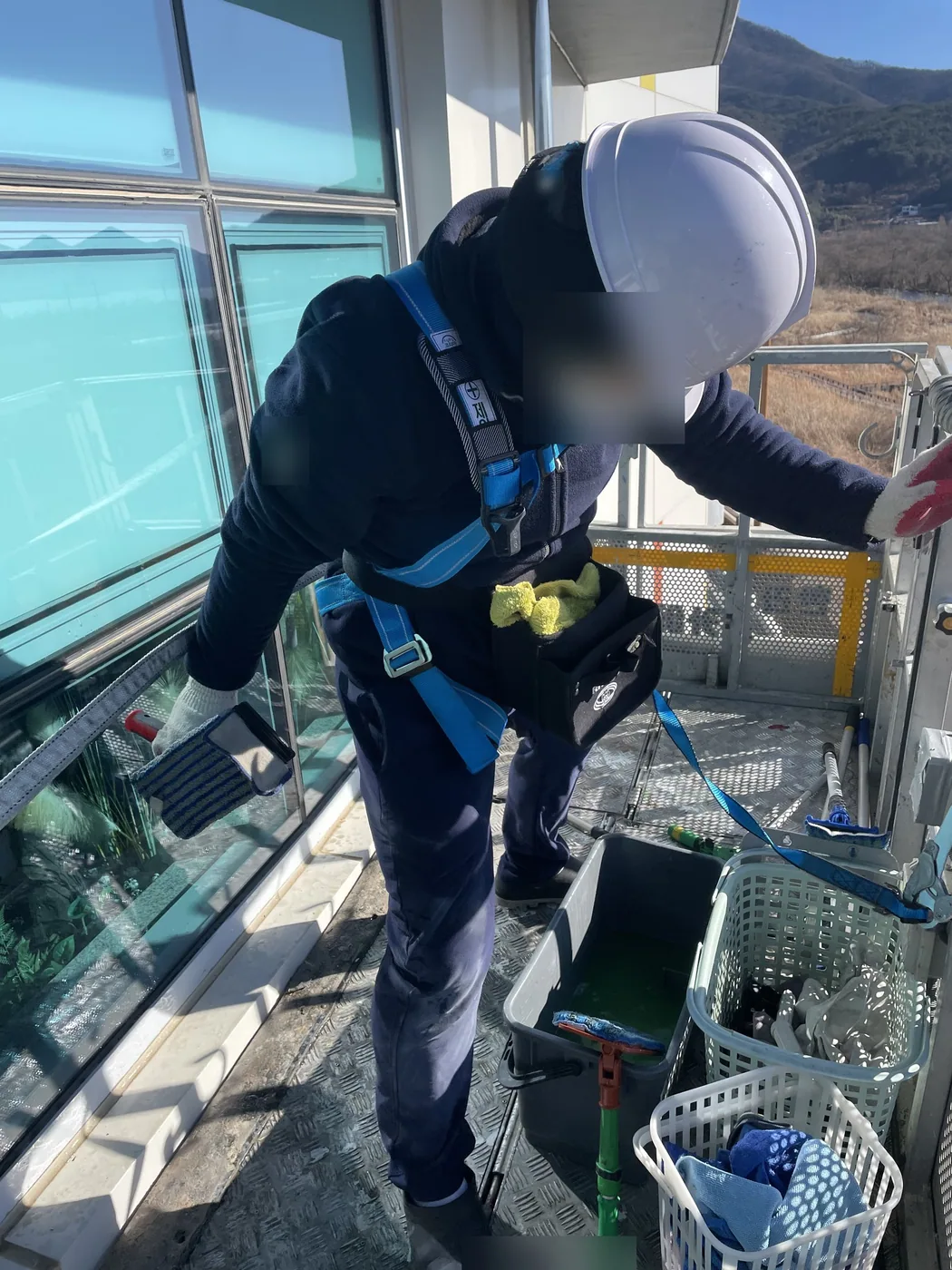 양산 교육시설 위층 곡면 창 앞 작업대에서 안전모와 안전대를 갖춘 작업자가 세척 패드로 창을 닦는 모습