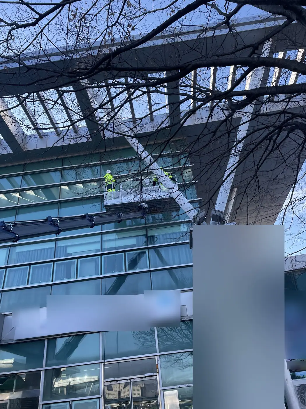 유리 외벽 윗단과 사선 기둥 사이로 올라간 고소작업차 바스켓에서 작업자 두 명이 작업하는 모습