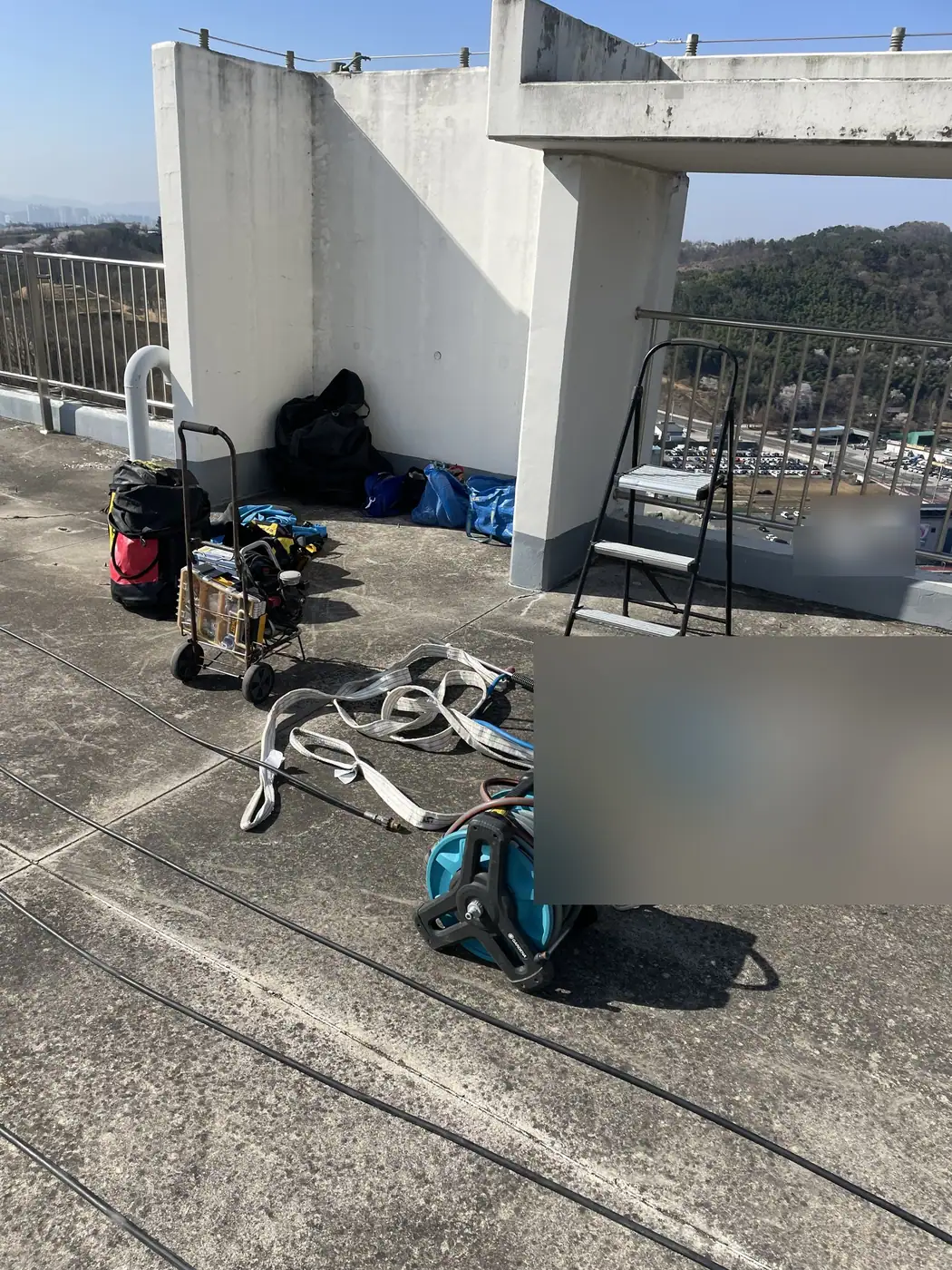 아파트 옥상 바닥에 펼쳐 둔 고정용 슬링과 물 호스 릴, 장비 가방과 카트, 난간 앞 접이식 사다리