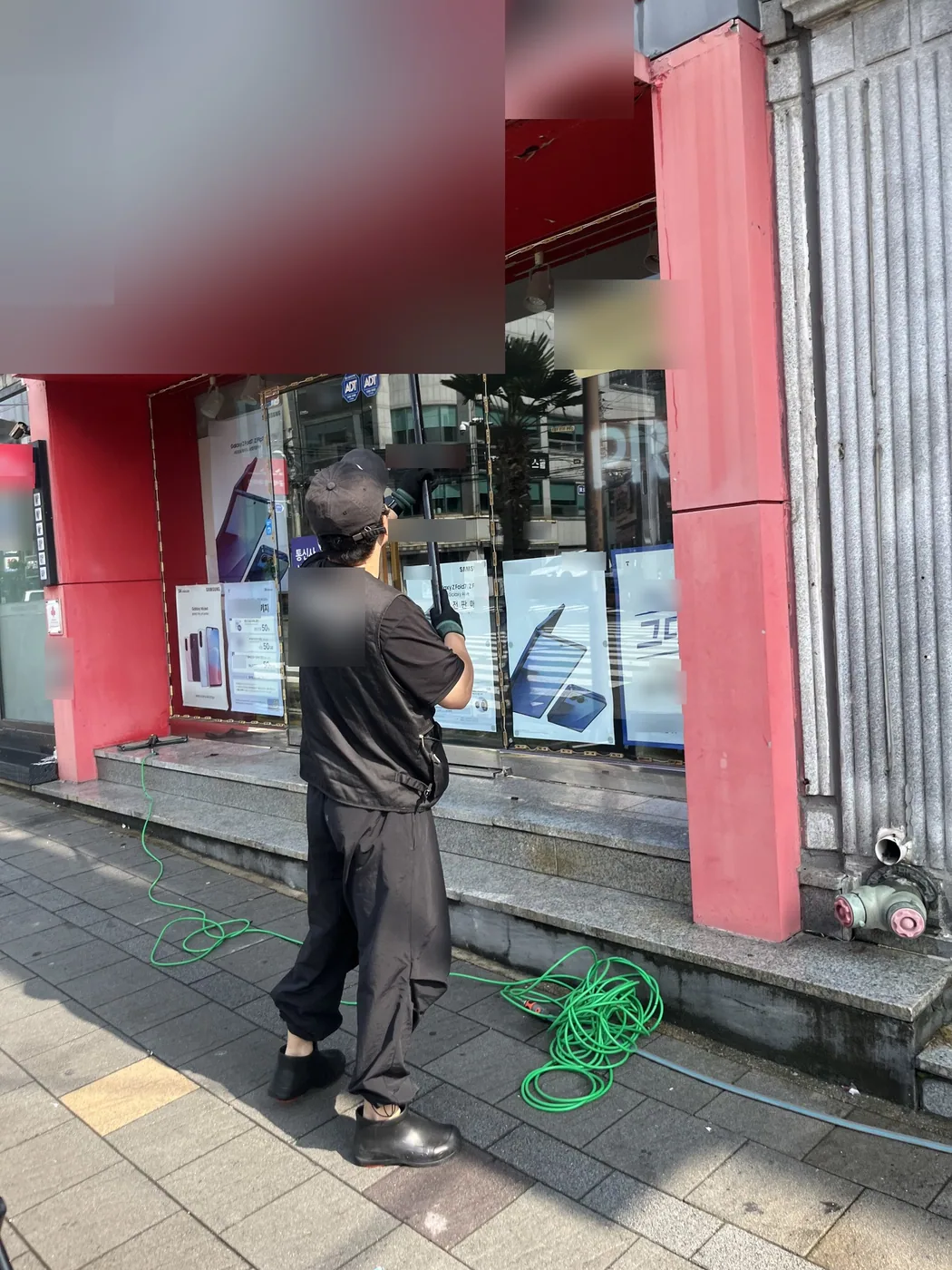 입구 앞 바닥에 호스를 풀어 둔 채 작업자가 폴대로 간판 판넬 위쪽을 닦는 뒷모습