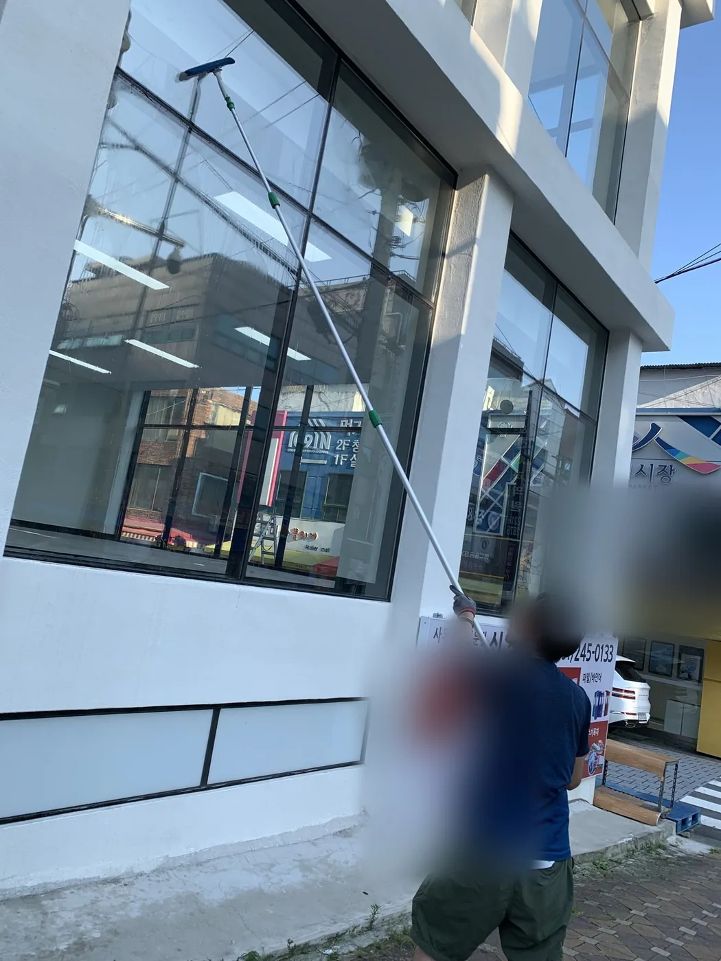 도로변 상가 유리면을 작업자가 폴대 도구로 밀어 세척하는 장면