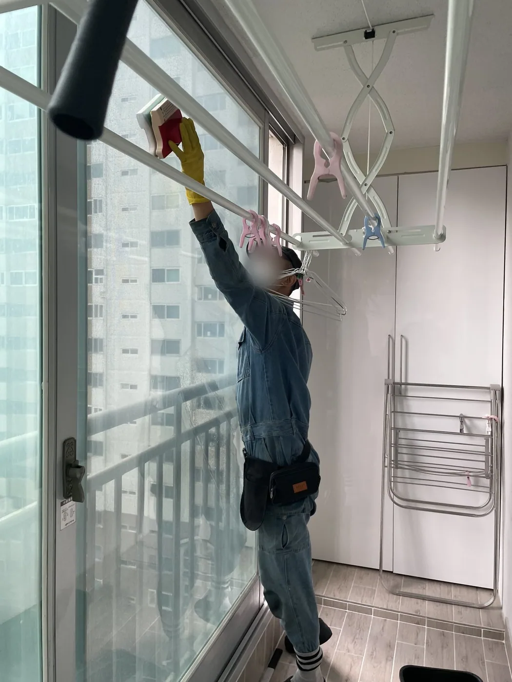작업자가 긴 폴대 도구로 베란다 창의 상단 고정 구획을 세척하는 장면