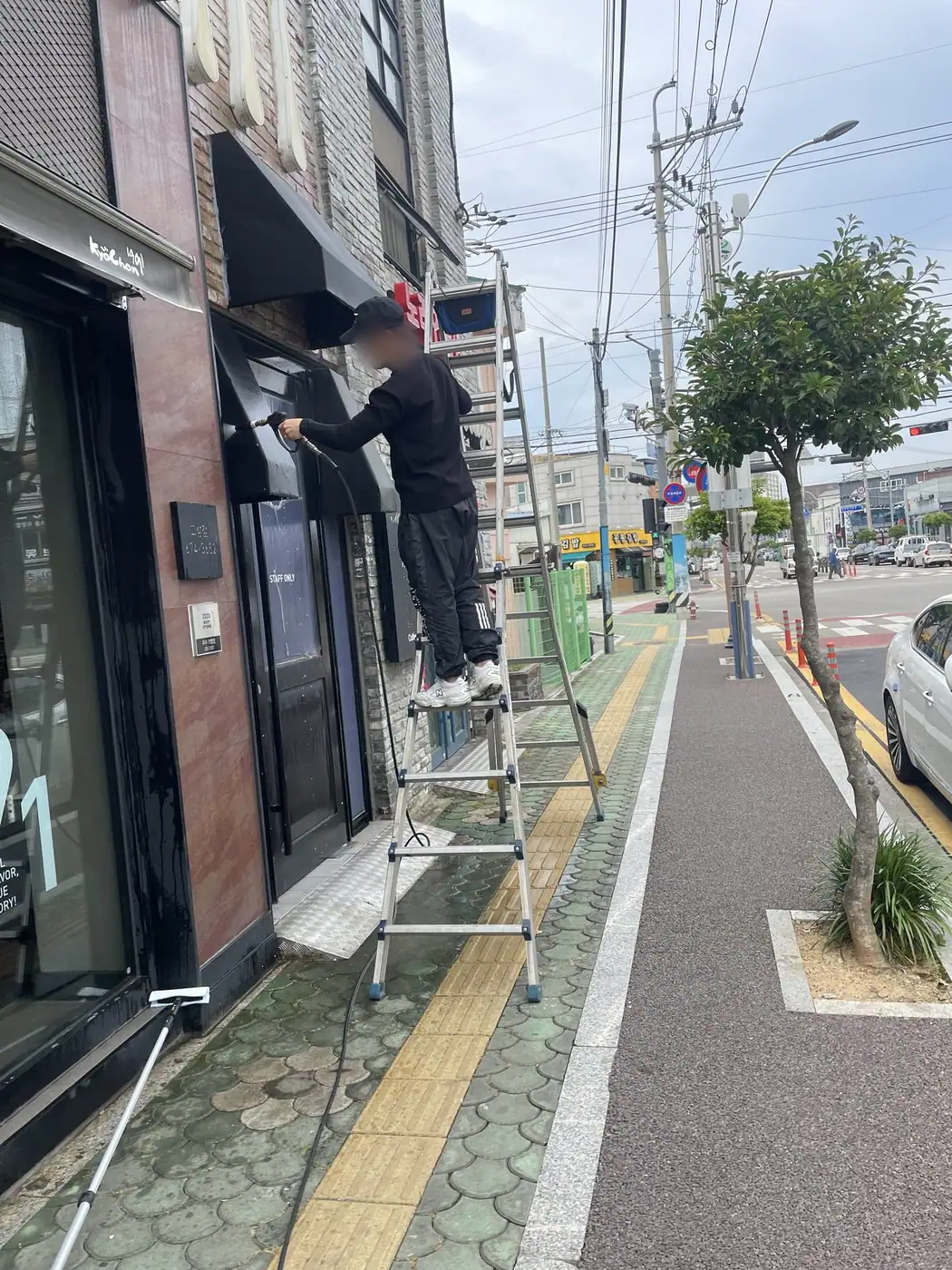 보도에 사다리를 세우고 작업자가 상가 어닝을 세척하는 전경 장면