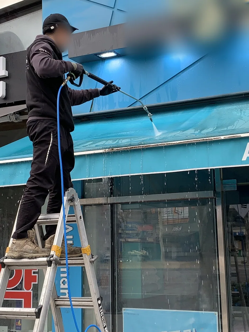 사다리에 올라선 작업자가 청록색 어닝 윗면을 세척하는 장면